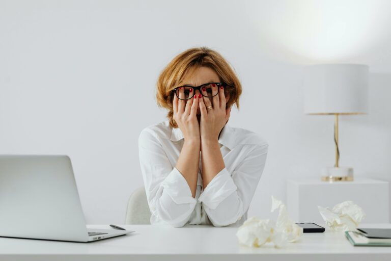 Une femme en chemise blanche, assise à son bureau, se prend la tête entre les mains avec un air épuisé et stressé. Devant elle, un ordinateur portable, un téléphone et des feuilles froissées témoignent d’un moment de frustration ou de surcharge de travail. L’arrière-plan épuré et lumineux contraste avec son expression de fatigue, illustrant le stress en milieu professionnel.