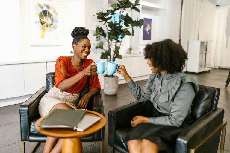 Deux collègues souriantes trinquent avec des tasses bleues dans un espace de travail moderne et convivial, illustrant un environnement professionnel chaleureux et détendu.